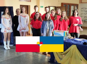 Grupa uczniów stoi przy punkcie szkolnej zbiórki charytatywnej, obok widać stół oraz flagi narodowe Ukrainy i Polski